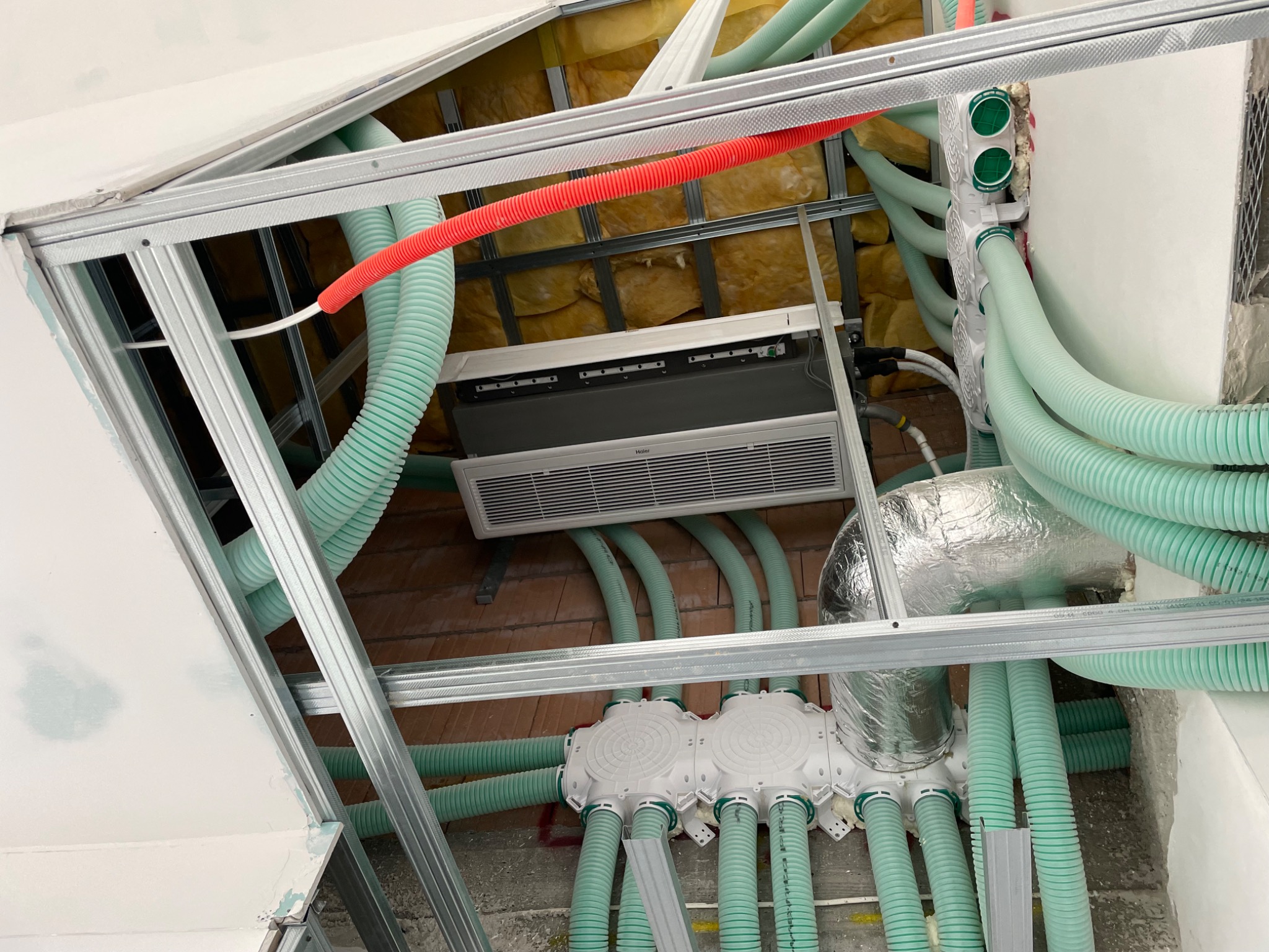 Widok z dołu na montaż sufitowy klimatyzacji Haier i system rekuperacji z licznymi elastycznymi przewodami wentylacyjnymi w budynku w trakcie wykończenia.