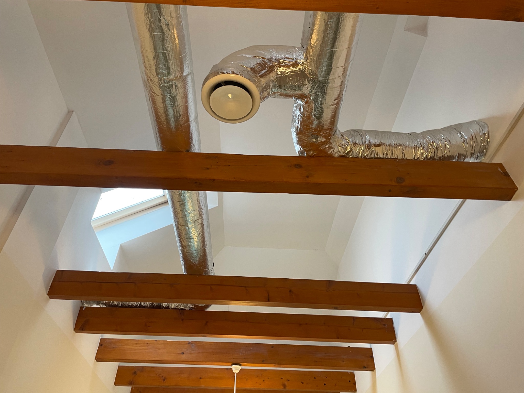 Widok z dołu na fragment instalacji wentylacyjnej z aluminiowymi przewodami i okrągłym nawiewnikiem zamontowanymi pod sufitem z drewnianymi belkami stropowymi.