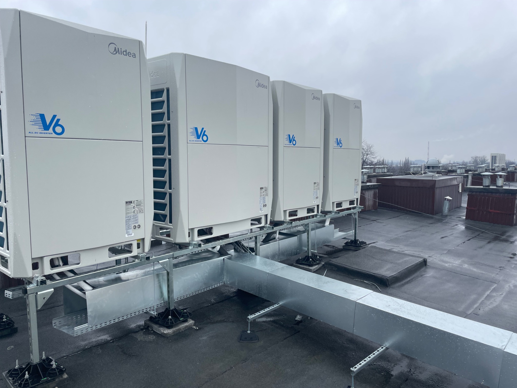 Cztery jednostki klimatyzacji Midea V6 zainstalowane na dachu budynku komercyjnego, połączone systemem kanałów wentylacyjnych, widok w pochmurny dzień.