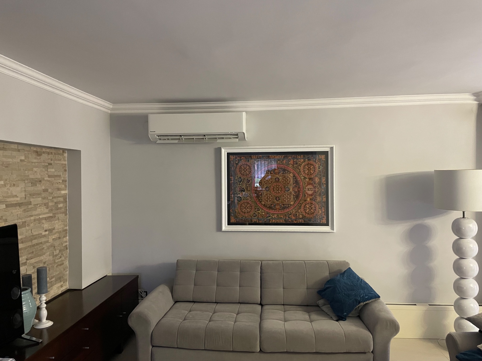 Biała jednostka klimatyzacyjna zamontowana nad obrazem w ramie, szara sofa z niebieską poduszką w salonie z lampą podłogową.