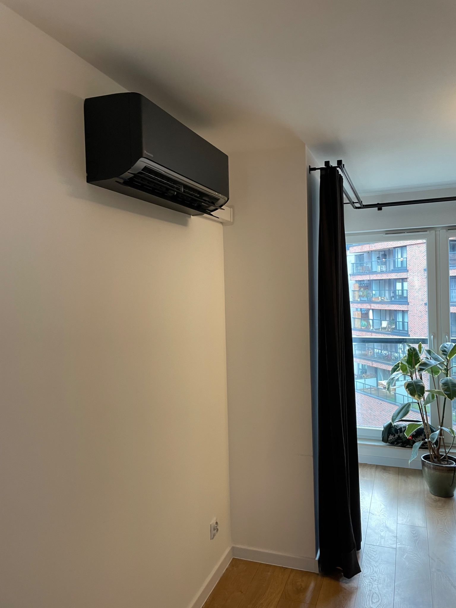 Czarny klimatyzator ścienny w nowoczesnym mieszkaniu, widok z boku, z zasłoniętym oknem i rośliną doniczkową w tle.