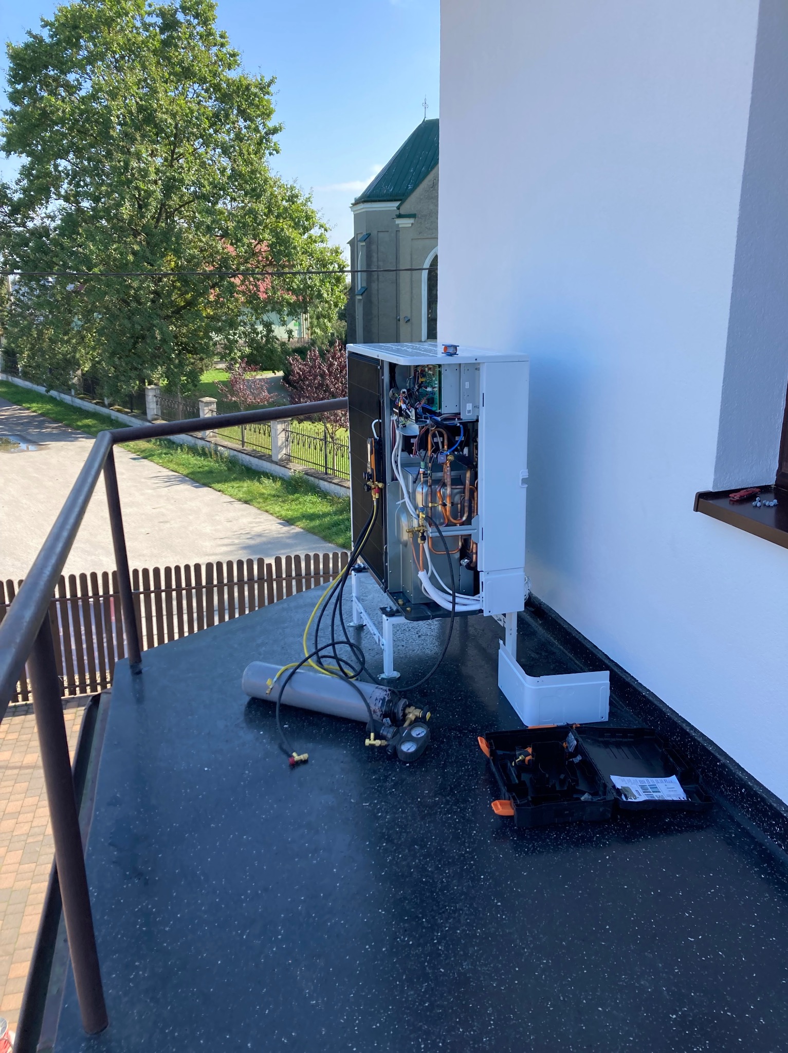 Biała jednostka zewnętrzna pompy ciepła podczas instalacji na balkonie, widoczne narzędzia i cylinder z gazem, w tle budynek kościoła i drzewa.