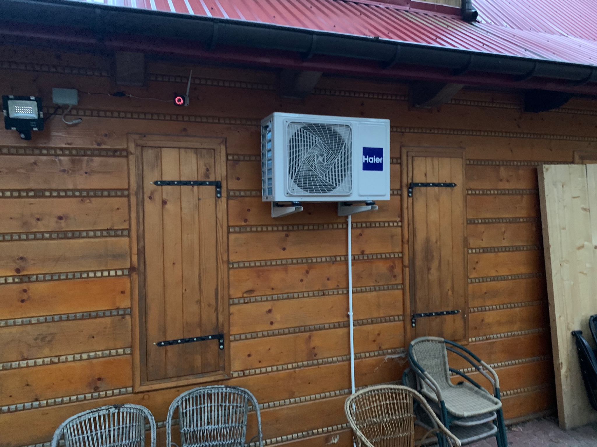 Biała jednostka zewnętrzna klimatyzatora Haier zamontowana na drewnianej ścianie budynku z okiennicami i widocznymi przewodami, oświetlona lampą zewnętrzną.