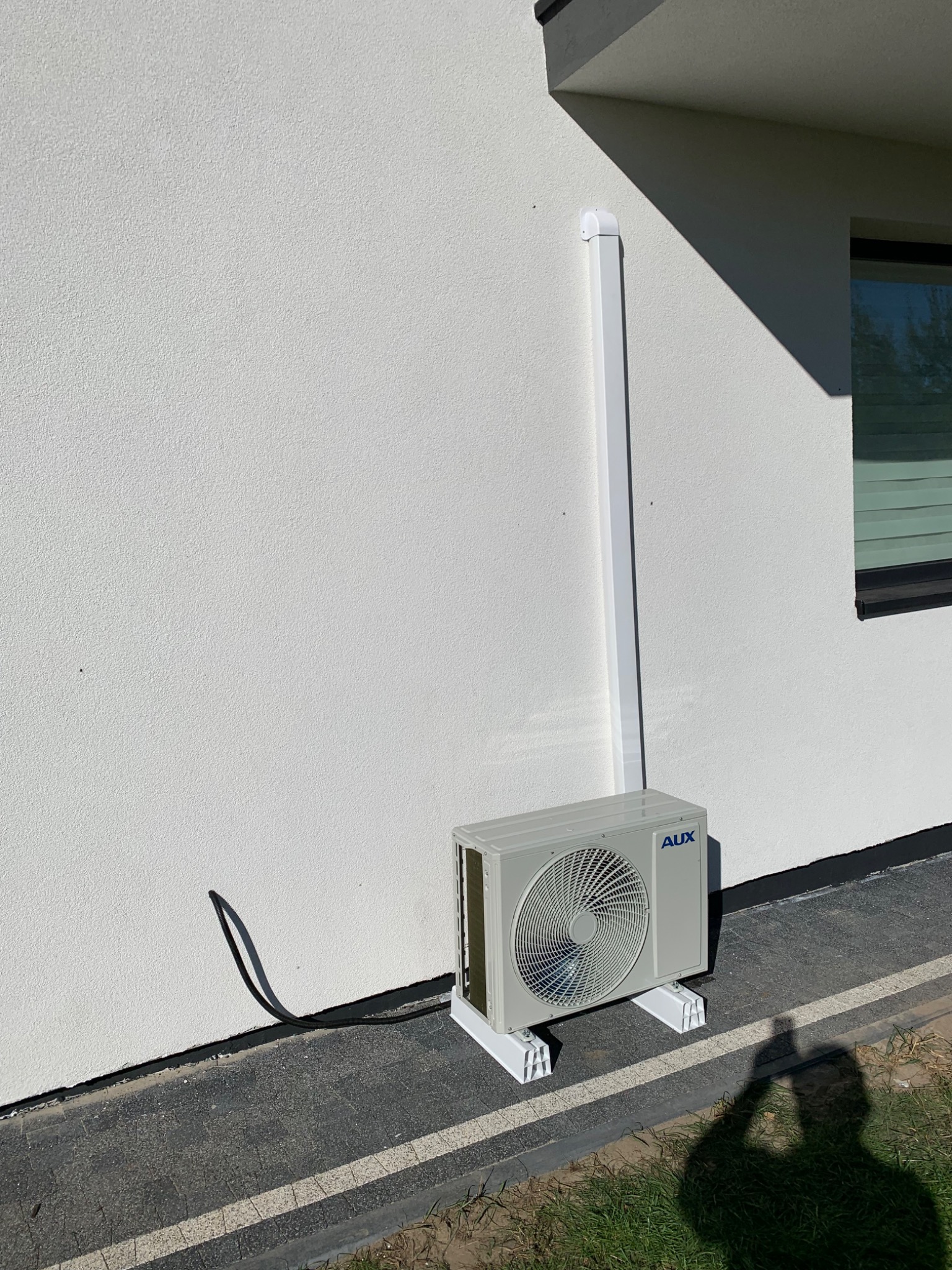 Zewnętrzna jednostka klimatyzacji AUX zamontowana na białej elewacji domu z widoczną instalacją odprowadzającą skropliny i cieniem osoby robiącej zdjęcie.