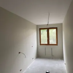 Pokój w trakcie przygotowania do malowania: ściany po szpachlowaniu i gruntowaniu, widoczne puszki elektryczne bez osprzętu, kabel zwisający z sufitu, puszka z farbą na surowej wylewce podłogowej...