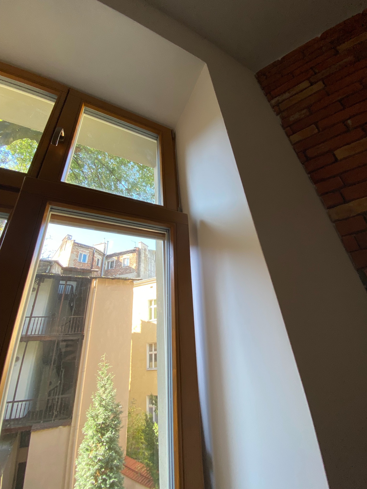 Pomalowana na biało wnęka okienna z drewnianą ramą okna, widok na podwórze kamienicy z zielenią i ceglaną ścianą w tle.