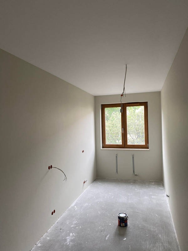 Pokój w trakcie przygotowania do malowania: ściany po szpachlowaniu i gruntowaniu, widoczne puszki elektryczne bez osprzętu, kabel zwisający z sufitu, puszka z farbą na surowej wylewce podłogowej...