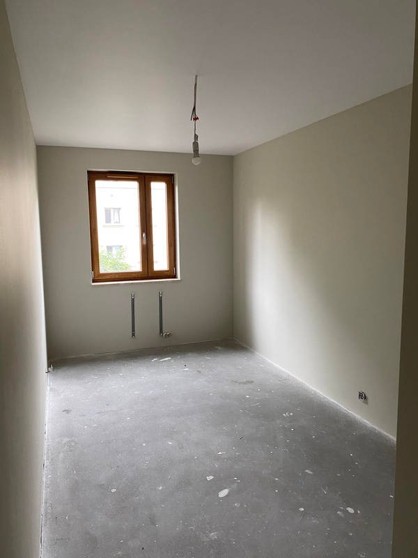 Pokój po szpachlowaniu i malowaniu na jasny kolor, widoczne instalacje grzewcze przy oknie i surowy beton na podłodze.