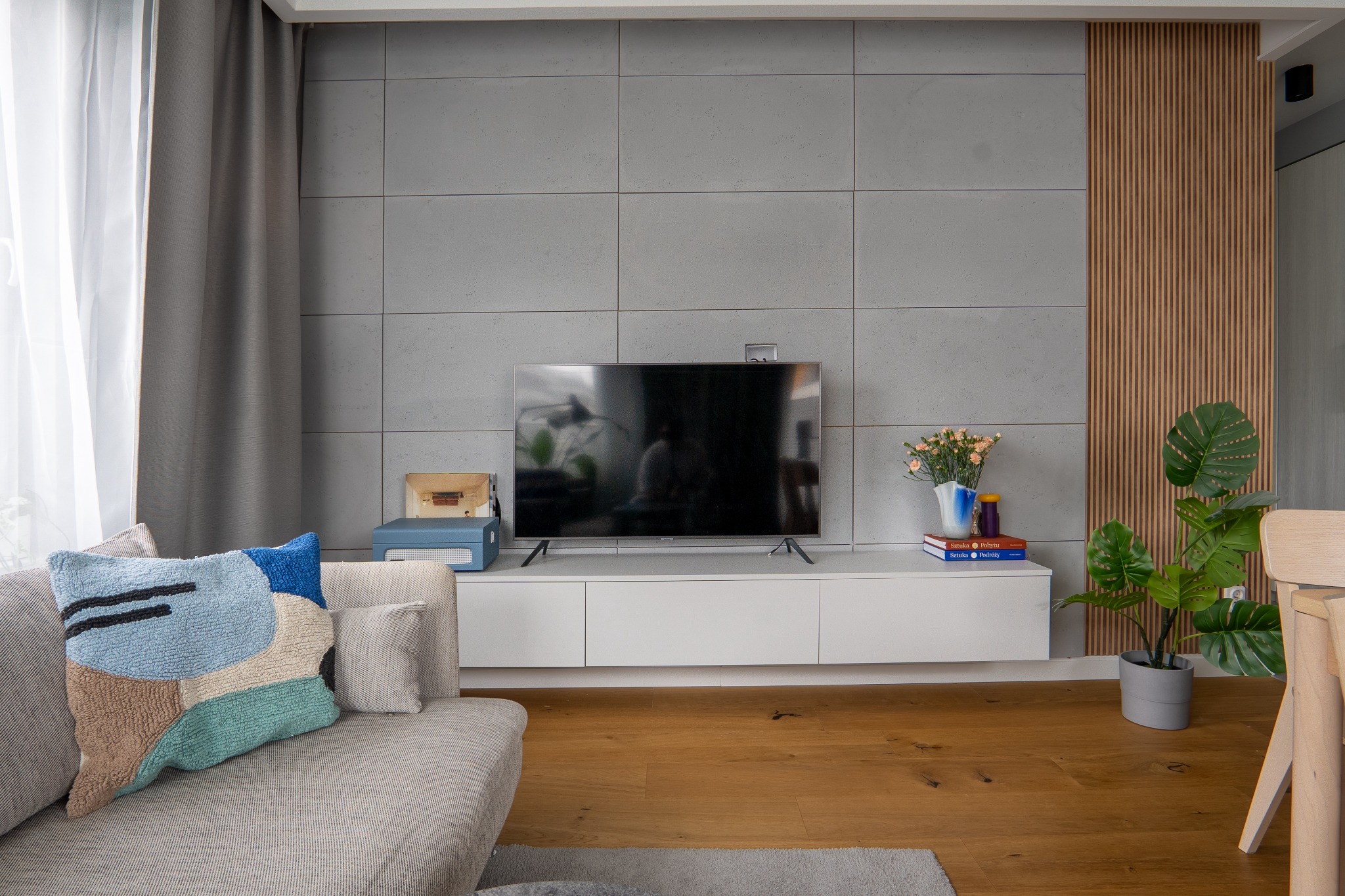 Nowoczesny salon z betonową ścianą, drewnianymi lamelami, białym regałem RTV, telewizorem, rośliną doniczkową i kanapą z dekoracyjną poduszką w odcieniach niebieskiego i brązu.