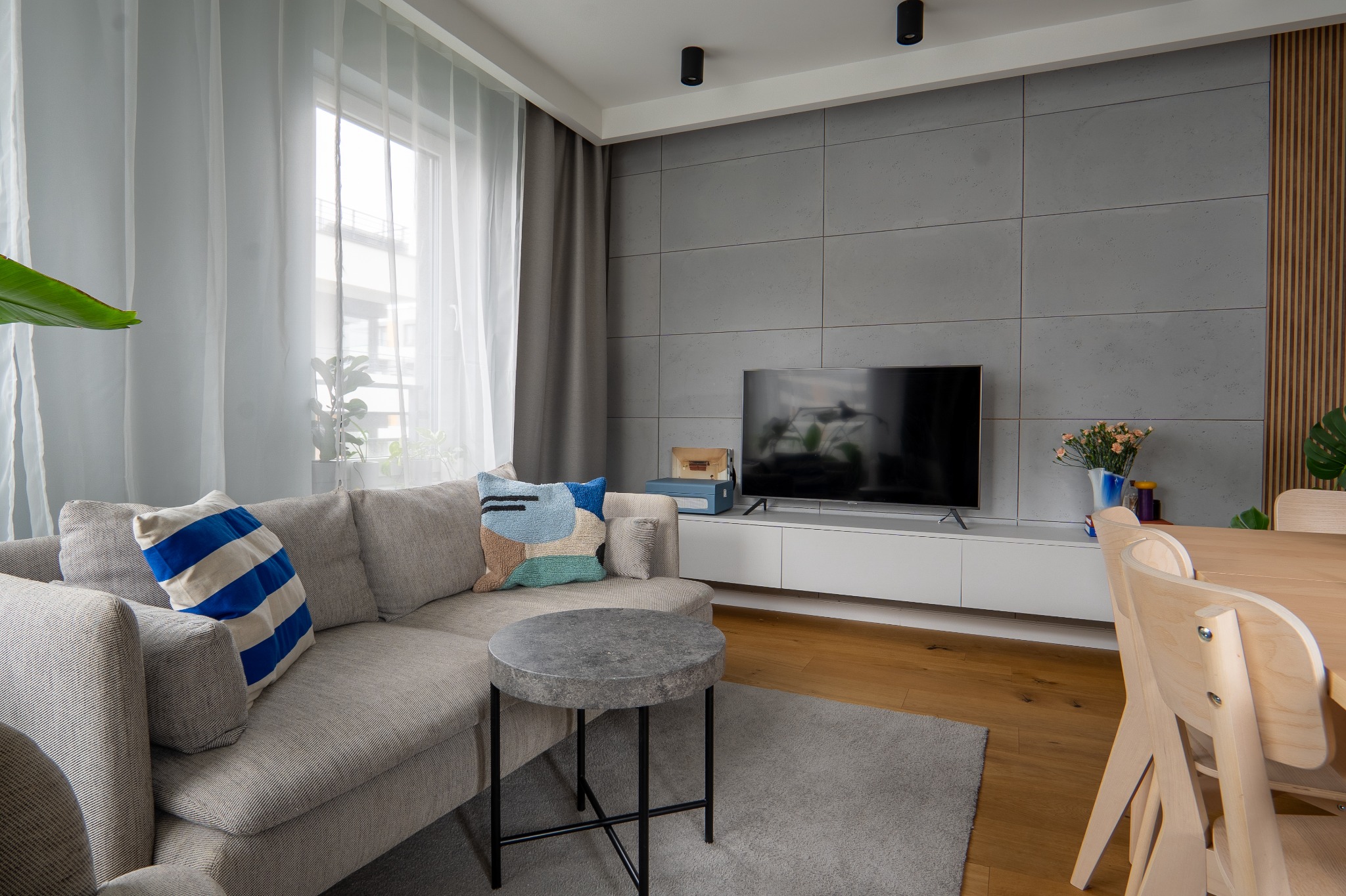 Jasny salon z szarą sofą, okrągłym stolikiem z szarym blatem, drewnianym stołem i krzesłami, szarą ścianą z telewizorem i białymi szafkami, zasłonami w oknie i dywanem na podłodze.