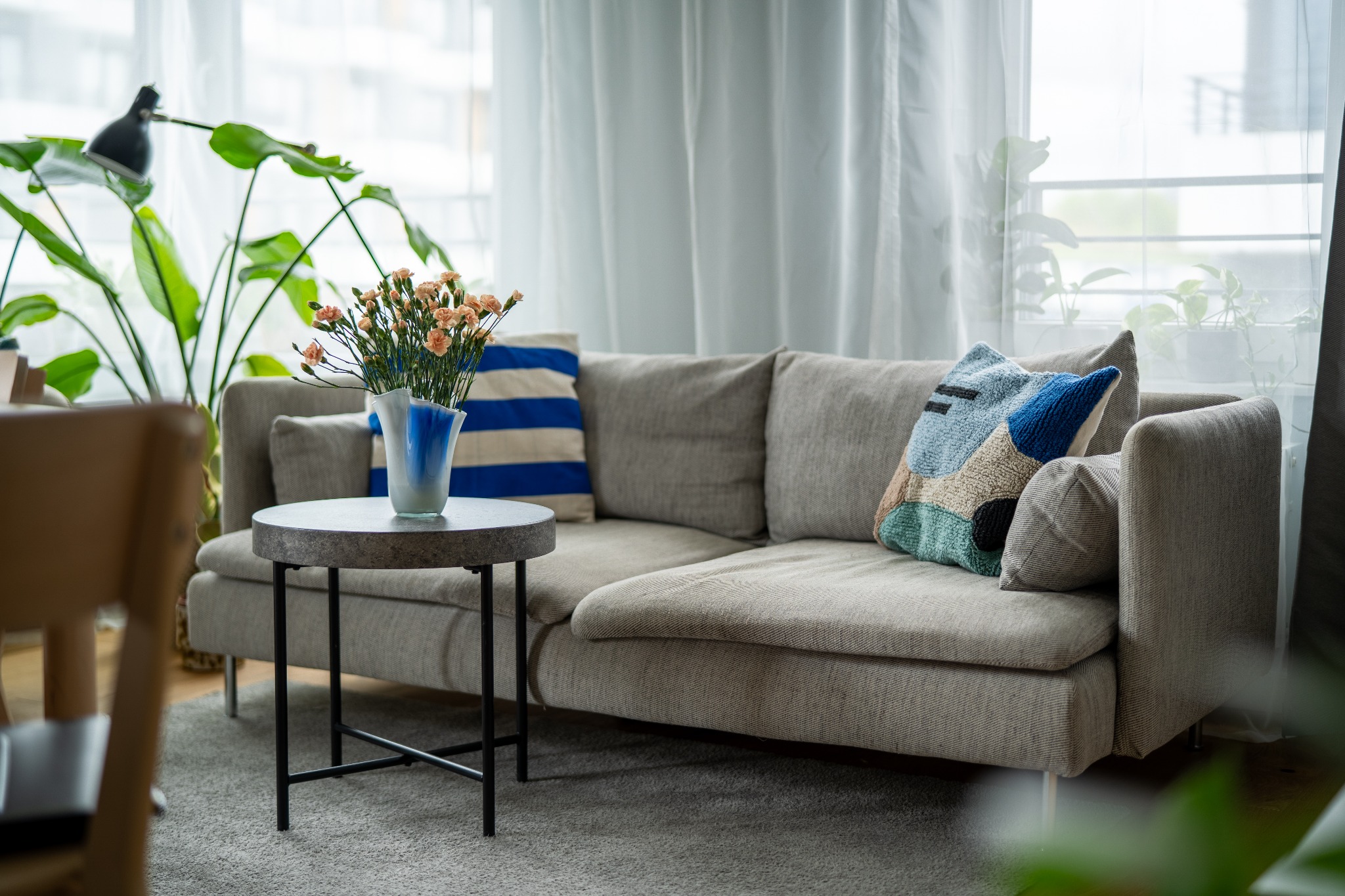 Jasnoszara sofa z poduszkami w paski i patchwork, okrągły stolik z kwiatami w niebieskim wazonie, za sofą widoczne okno z białymi zasłonami i roślina doniczkowa.