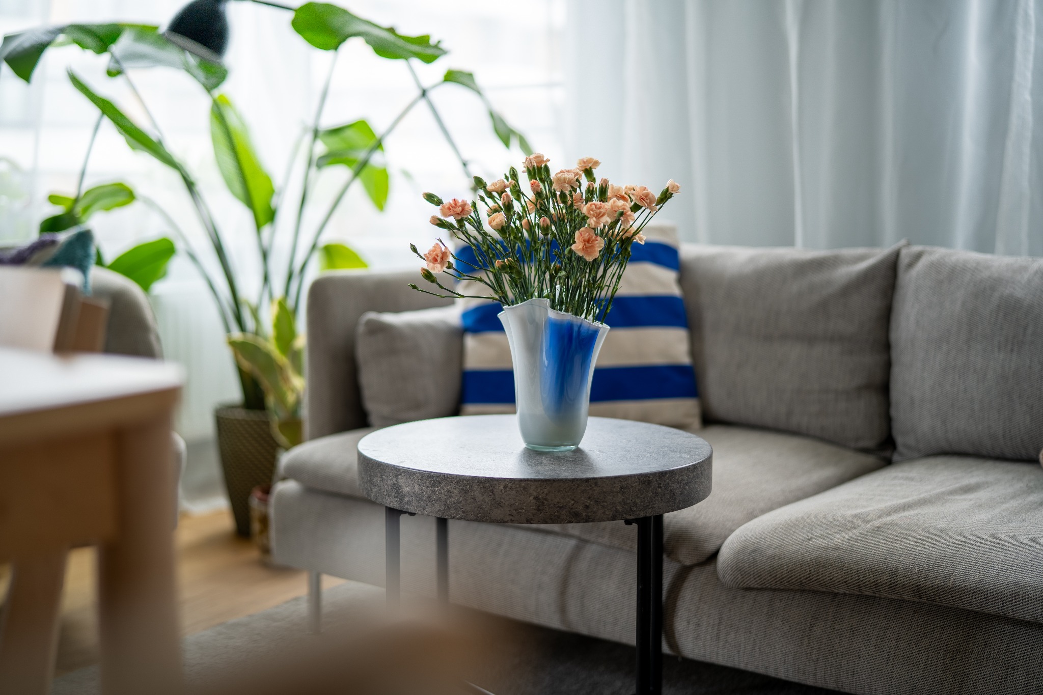 Bukiet bladoróżowych goździków w białym wazonie z niebieskimi akcentami, stojący na okrągłym, szarym stoliku kawowym na tle szarej sofy z poduszką w niebiesko-białe paski, całość dopełnia duża...