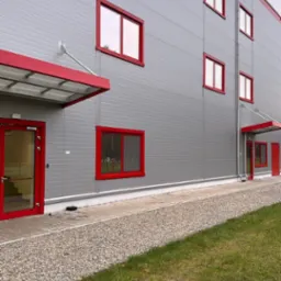 Zewnętrzny widok hali z szarych płyt warstwowych z czerwonymi obramowaniami okien i drzwi, widoczny chodnik z kamienia i trawnik.