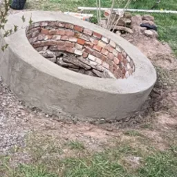 Owalna, murowana konstrukcja z cegieł, częściowo otynkowana szarym cementem, otoczona ziemią i rosnącą trawą, prawdopodobnie w trakcie budowy, widoczne narzędzia w tle.