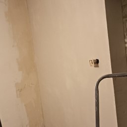 nie mam firmy - Wnętrze w trakcie prac wykończeniowych: ściany po nałożeniu warstwy szpachli, widoczne puszki elektryczne, narzędzia pracy (wiertarka, paca) oraz worek z gipsem.