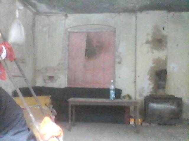 Wnętrze remontowanego pomieszczenia z widocznymi śladami po pracach na ścianach, z centralnie umieszczonym otworem drzwiowym pomalowanym na różowo, z widocznymi meblami i butelką wody na stole...