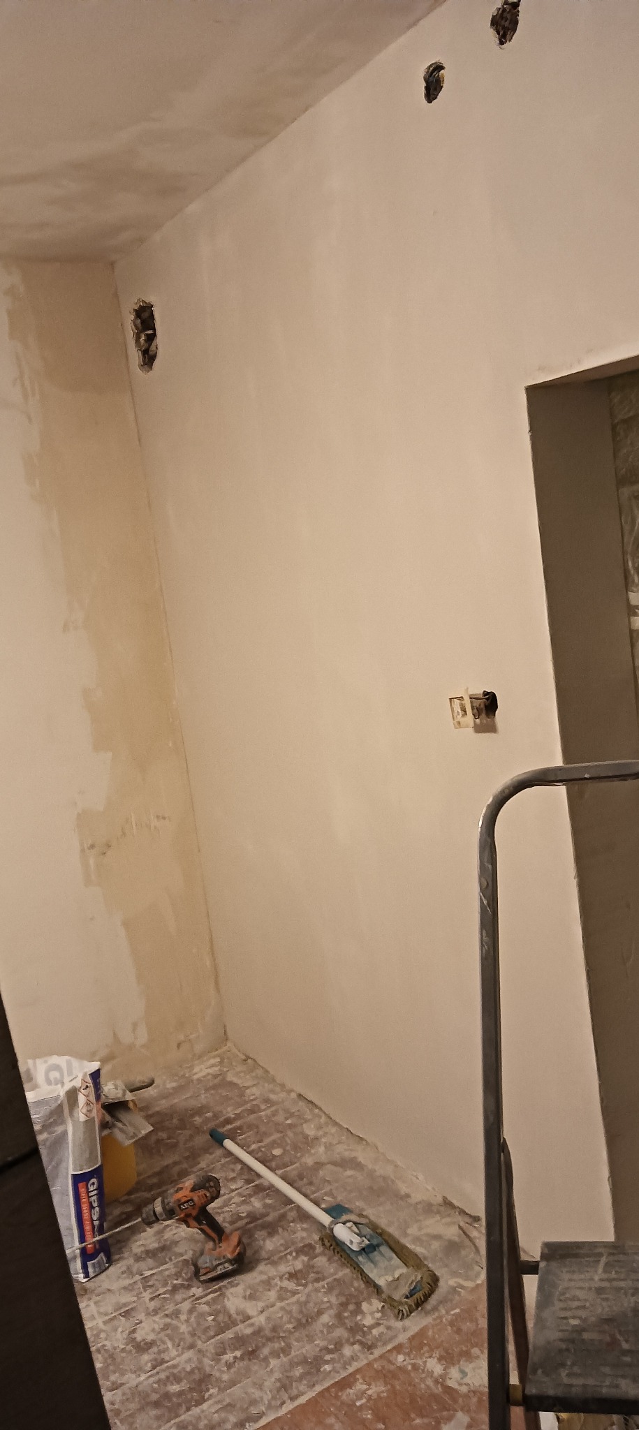 Wnętrze w trakcie prac wykończeniowych: ściany po nałożeniu warstwy szpachli, widoczne puszki elektryczne, narzędzia pracy (wiertarka, paca) oraz worek z gipsem.