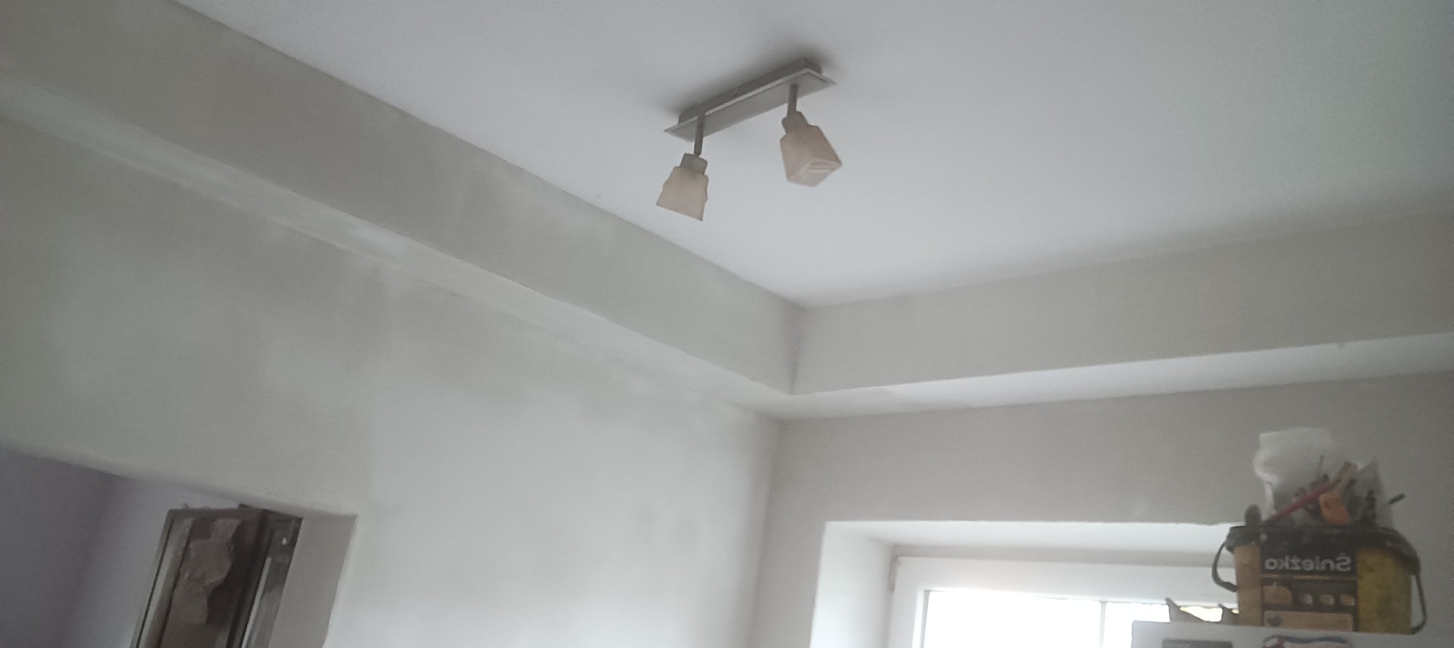 Świeżo pomalowane na biało wnętrze pokoju z widoczną lampą sufitową, listwami przysufitowymi i fragmentem okna z narzędziami malarskimi na parapecie.