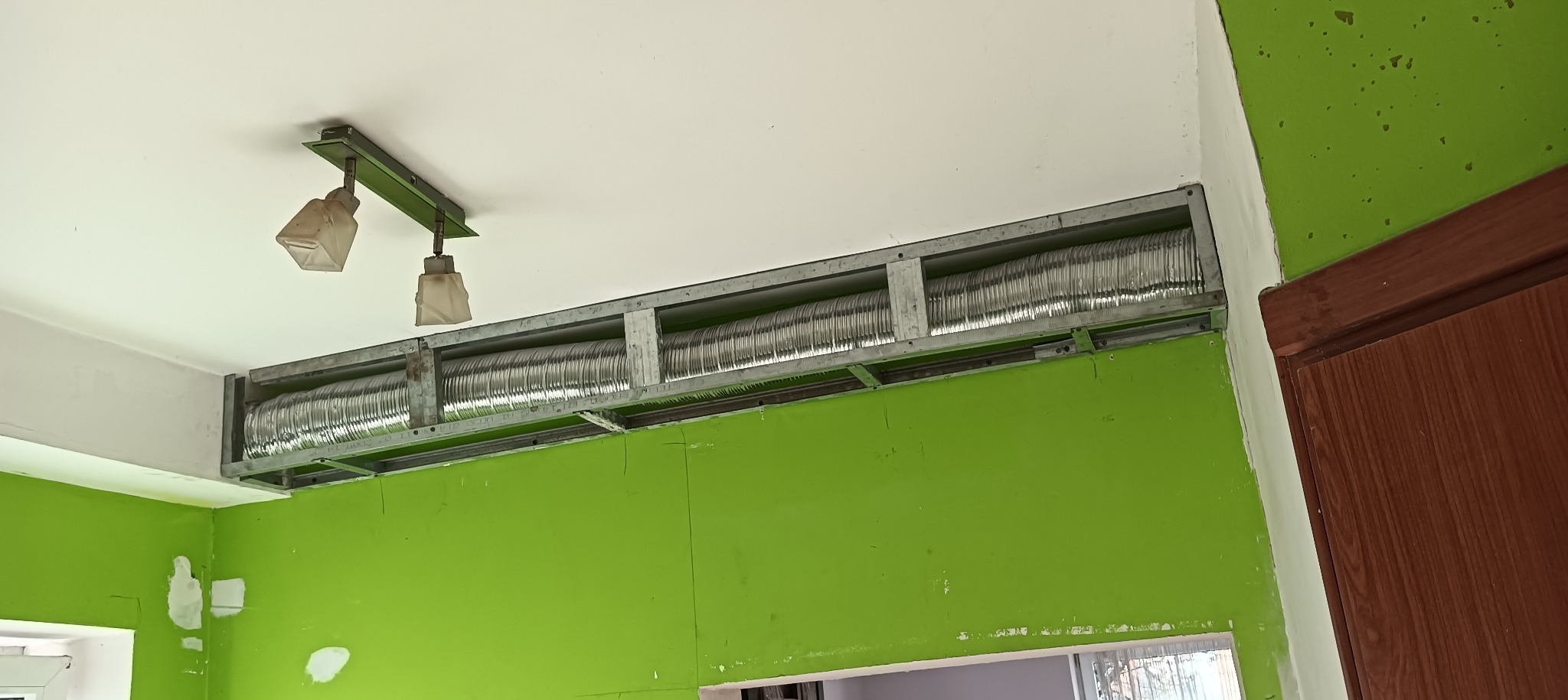 Metalowa konstrukcja pod sufitem z widoczną karbowaną rurą wentylacyjną, zielona ściana i stara lampa na suficie.