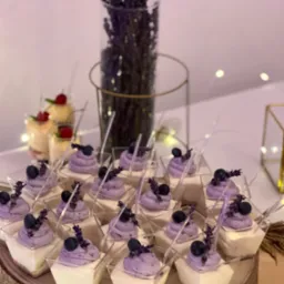 Deserki w małych, kwadratowych naczyniach na drewnianej podstawie. Każdy deser składa się z białej warstwy na dole, fioletowej pianki na wierzchu, borówki i gałązki lawendy. W tle wazon z lawendą...