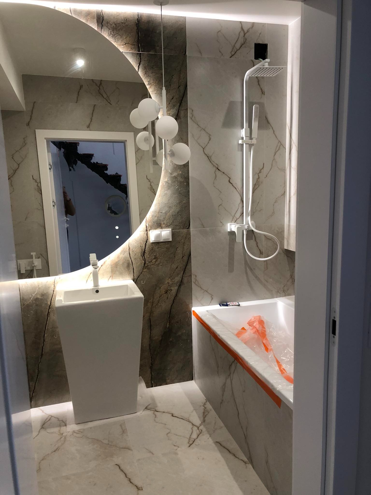 Nowoczesna łazienka z marmurowymi płytkami, owalnym lustrem z podświetleniem LED, wolnostojącą umywalką, białą armaturą i wanną zabezpieczoną folią ochronną podczas remontu.