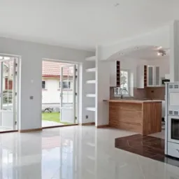 Jasne wnętrze domu w Ełku z otwartą kuchnią, białymi ścianami, błyszczącą podłogą i piecem wolnostojącym. Widok na ogród przez otwarte dwuskrzydłowe drzwi balkonowe.