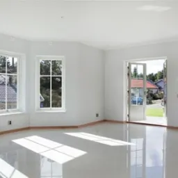 Jasne, puste wnętrze domu z białymi ścianami, dużymi oknami z widokiem na zieleń i błyszczącą, jasną podłogą z widocznymi promieniami słońca.