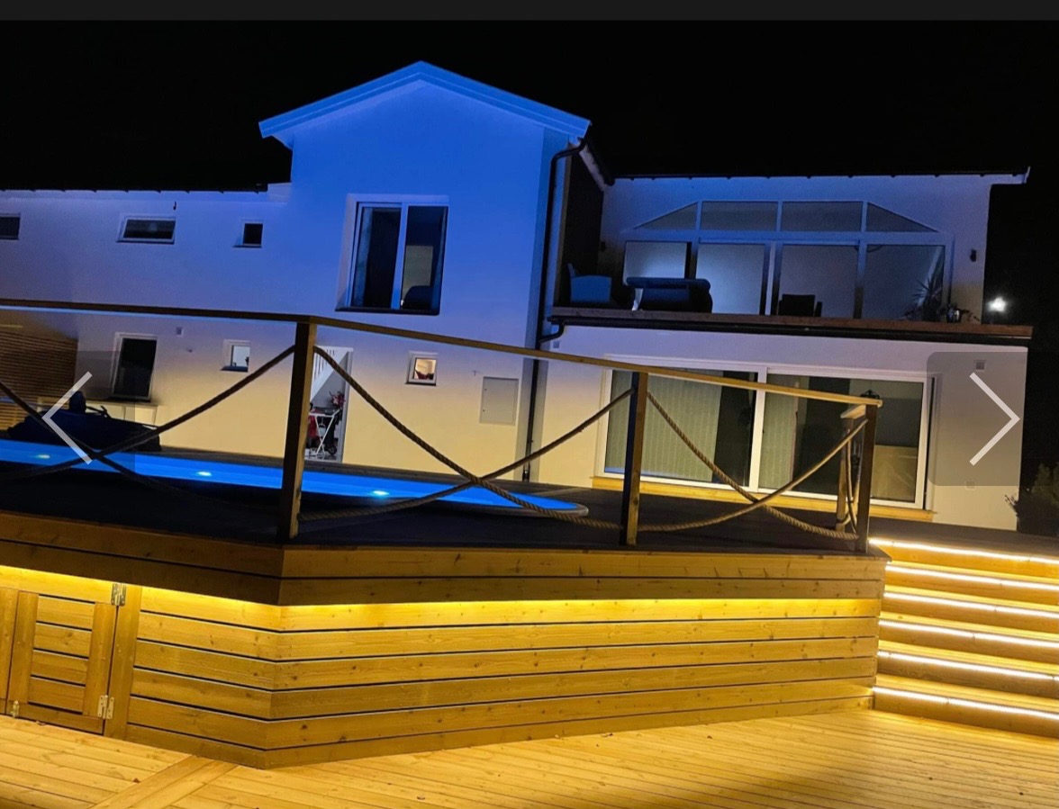 Nowoczesny dom z podświetlonym basenem i drewnianym tarasem, oświetlenie LED podkreśla architekturę w nocnej scenerii.