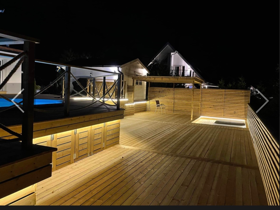 Oświetlony taras z drewna z zabudową pod podestem, widoczny basen i dom w tle, nocna sceneria.