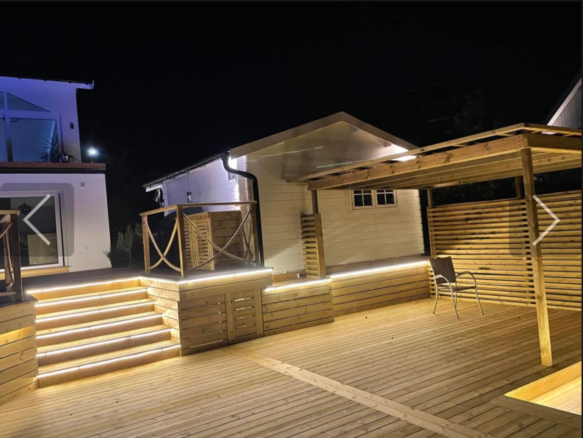 Oświetlony drewniany taras przy białym domu z drewnianą pergolą i schodami z podświetleniem LED, nocne ujęcie.