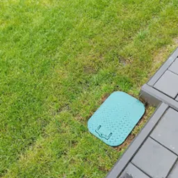 Zielony właz inspekcyjny ukryty w trawniku, obok ułożona szara kostka brukowa.