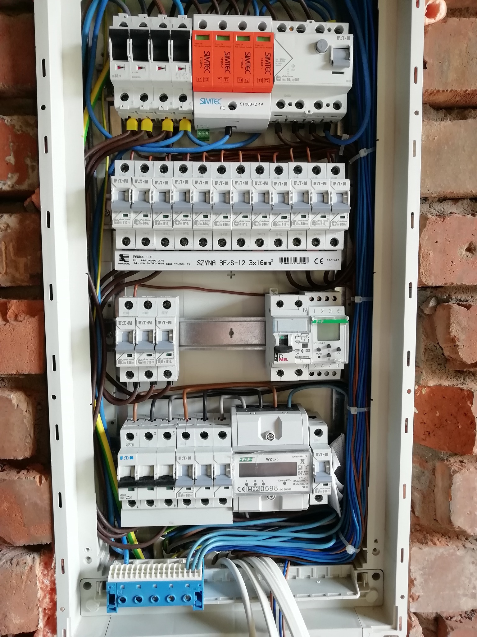 Rozdzielnica elektryczna z zainstalowanymi bezpiecznikami, wyłącznikami i przewodami, zamontowana na ceglanej ścianie, widoczne elementy firm Eaton, Pawbol i Fael.