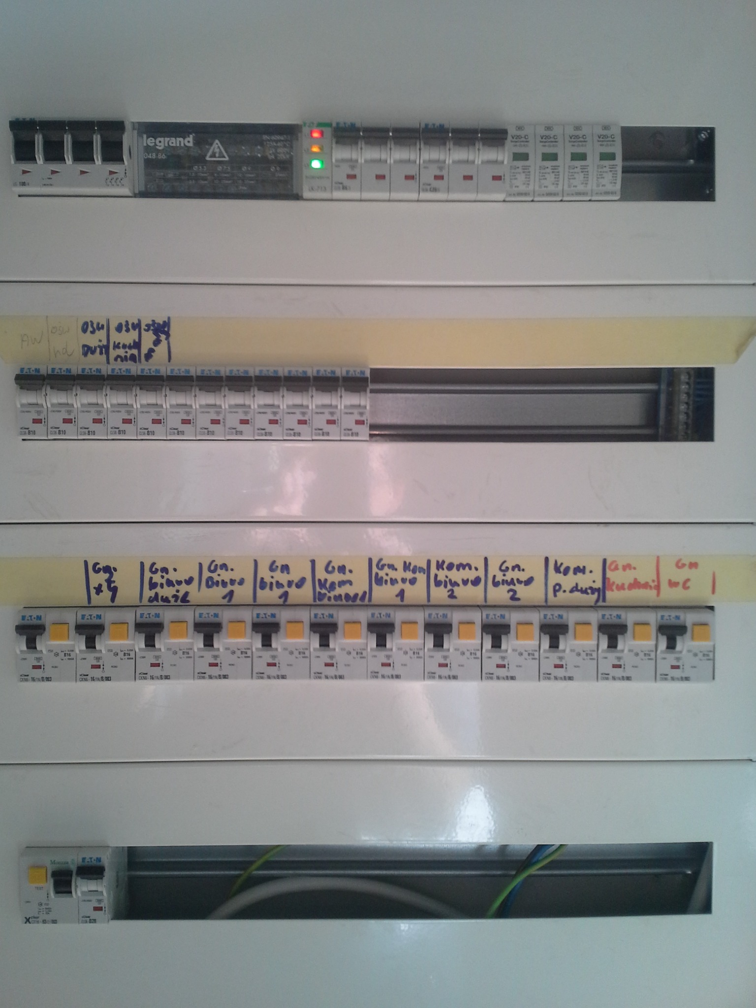 Rozdzielnica elektryczna z widocznymi bezpiecznikami różnych producentów (Legrand, Eaton), opisana taśmą z odręcznymi notatkami identyfikującymi obwody.