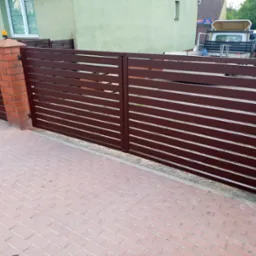 Nowoczesna brama wjazdowa z poziomych, brązowych profili, zamontowana na ceglanych słupkach z kostką brukową na podjeździe.