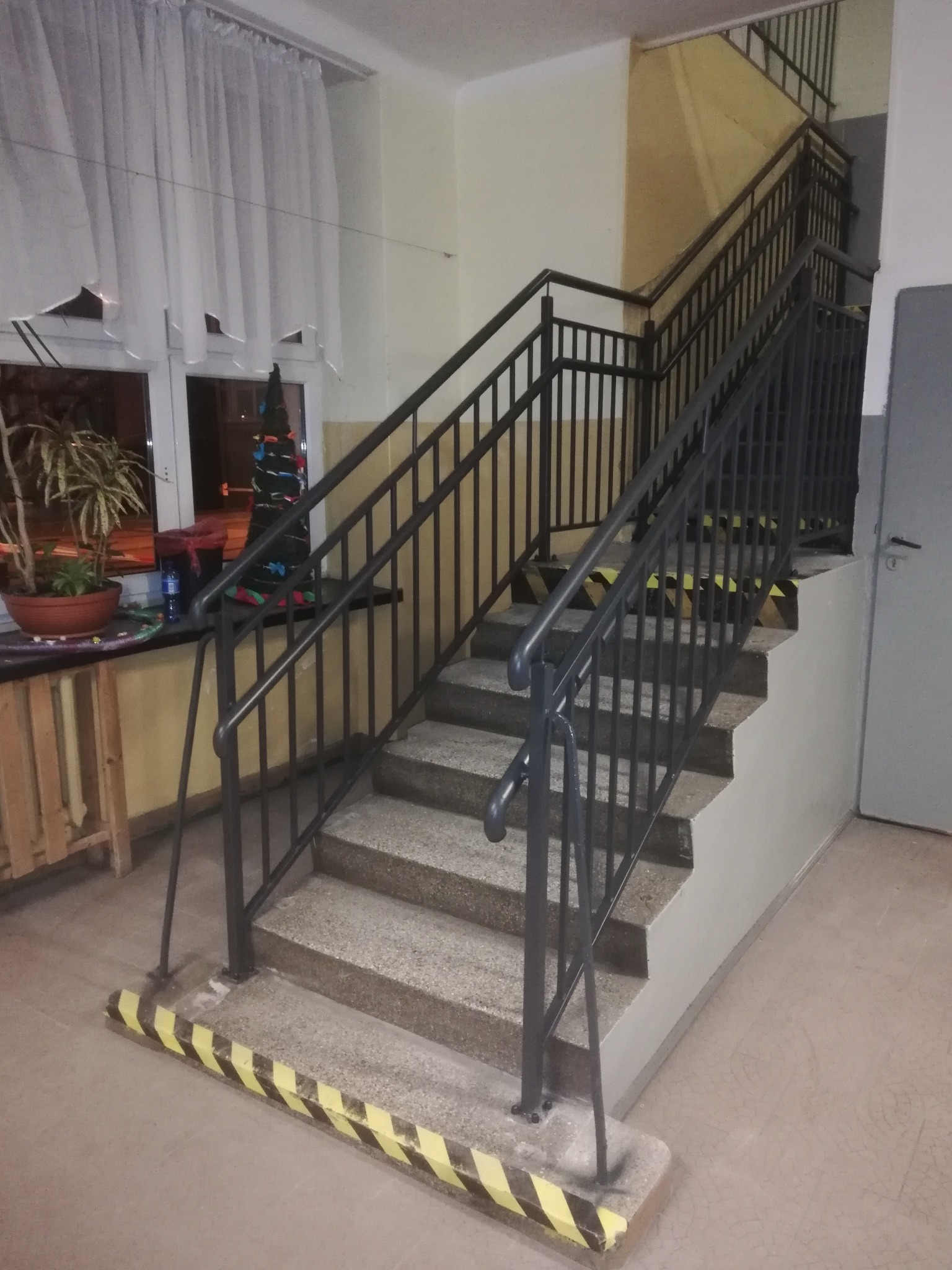 Wewnętrzne schody z metalową balustradą, zabezpieczone taśmą ostrzegawczą na krawędziach stopni, widok z dołu.