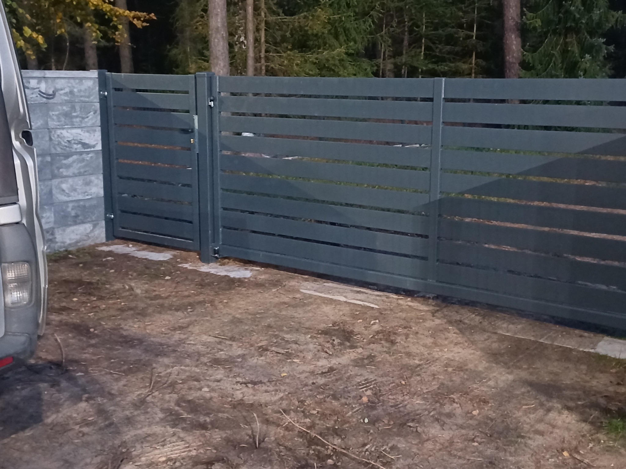 Nowoczesne ogrodzenie metalowe z poziomymi panelami, przylegające do betonowego słupka i częściowo zasłonięte przez samochód dostawczy, na tle leśnym.