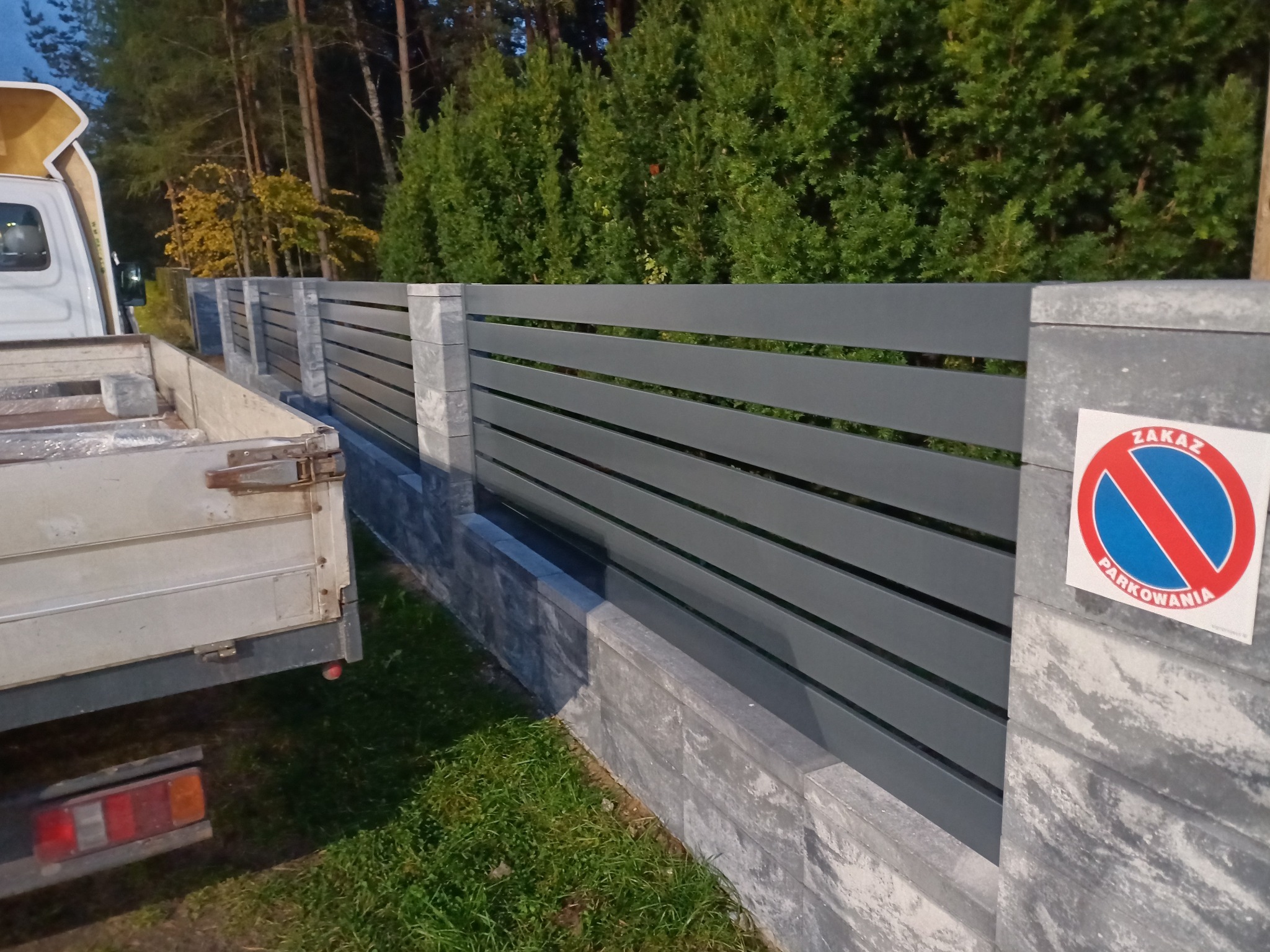 Nowoczesne ogrodzenie panelowe z szarego metalu i betonowych słupków, z naklejką 'Zakaz Parkowania', częściowo zasłonięte przez samochód dostawczy