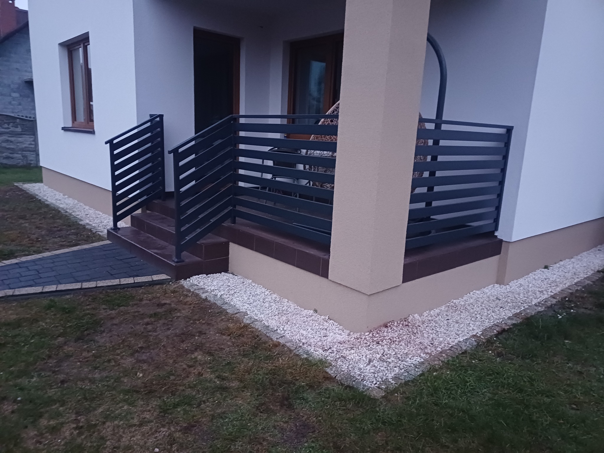 Nowoczesne, poziome, metalowe balustrady w kolorze grafitowym prowadzące do wejścia domu z kamienną ścieżką i ozdobnymi kamykami.