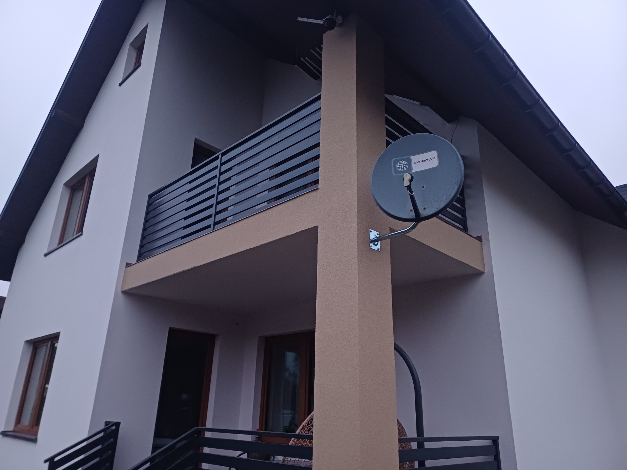 Nowoczesny dom z balkonem zabezpieczonym czarną, poziomą balustradą metalową, z anteną satelitarną zamontowaną na kolumnie podtrzymującej balkon, widok z dołu.