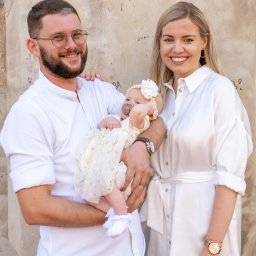 Julka Klimczak - Rodzinne zdjęcie: Uśmiechnięci rodzice w białych strojach trzymają na rękach niemowlę w kremowej sukience z kokardą, na tle ściany z delikatnymi napisami.