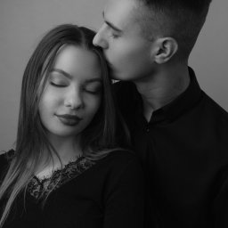 Julka Klimczak - Czarno-białe zbliżenie: mężczyzna w czarnej koszuli całuje kobietę w czoło, oboje z zamkniętymi oczami na jednolitym tle.