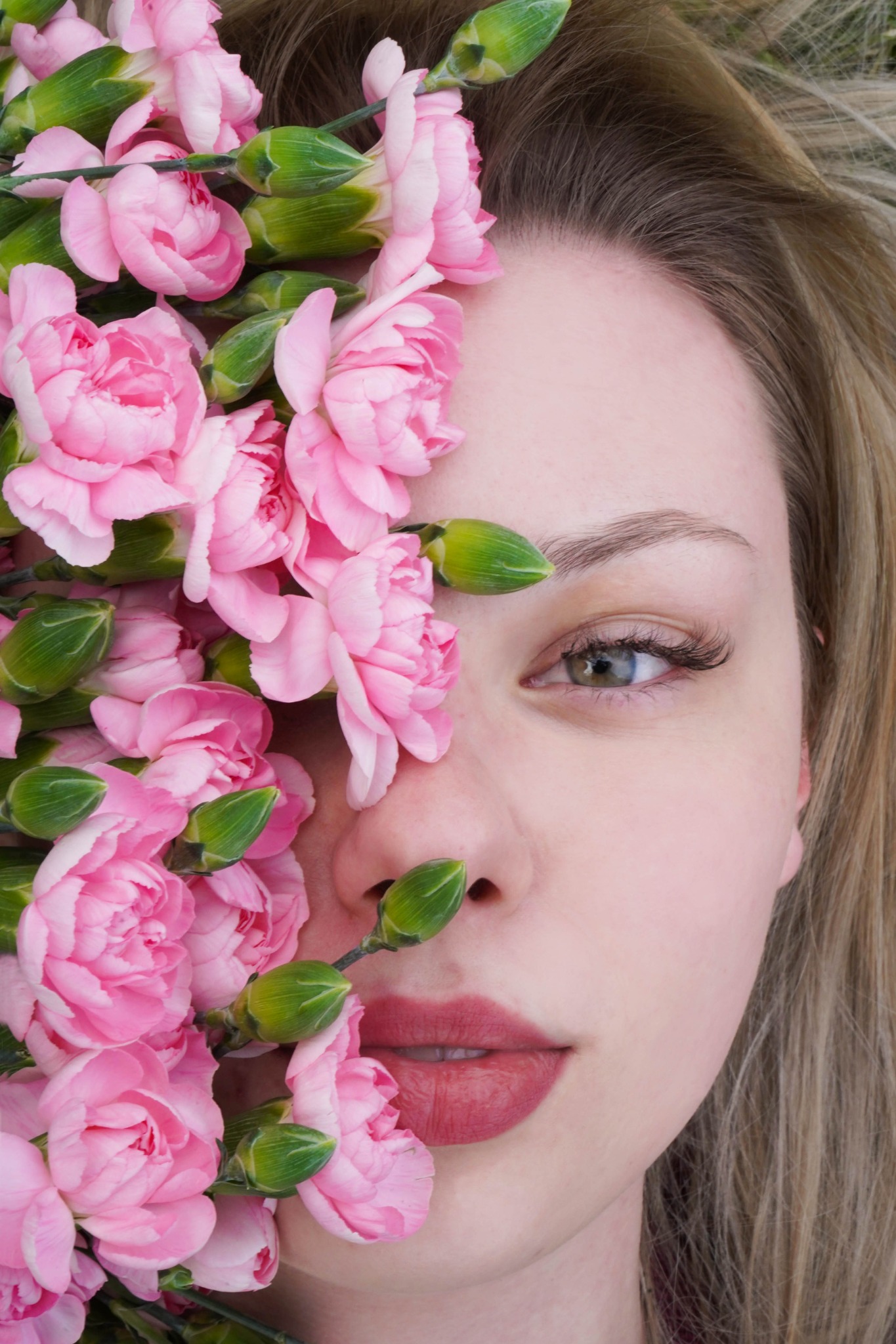 Portret kobiety z połową twarzy zasłoniętą przez bukiet różowych goździków, makijaż z podkreślonymi rzęsami i ustami.