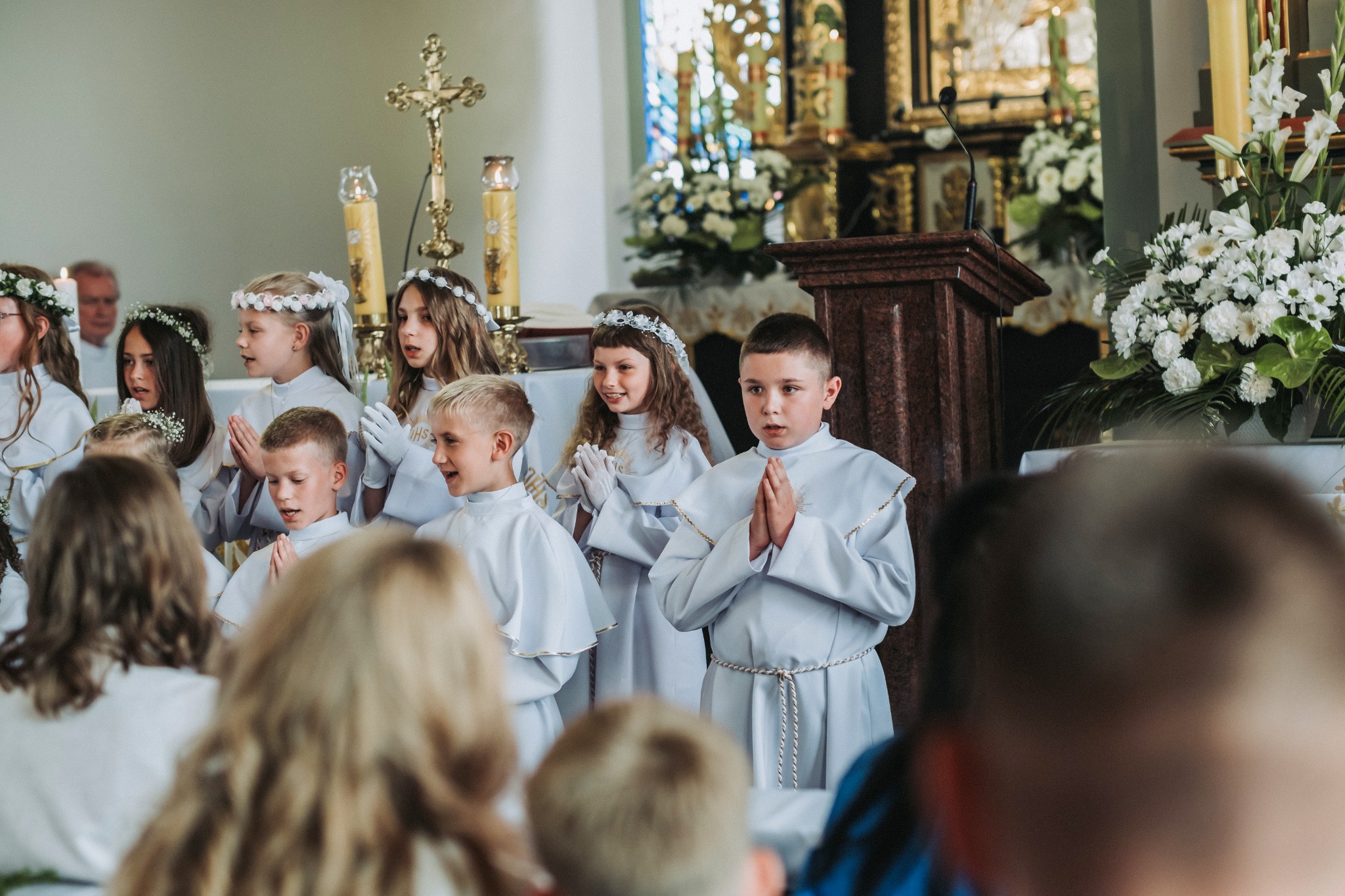 Dzieci w białych strojach komunijnych, z rękami złożonymi do modlitwy, stoją w kościele na tle ołtarza i dekoracji kwiatowych.