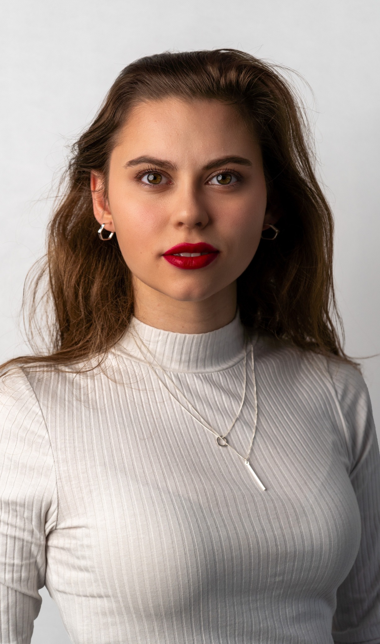 Portret kobiety z długimi brązowymi włosami, w białej bluzce z golfem, z srebrną biżuterią i czerwonymi ustami, na jasnym tle.