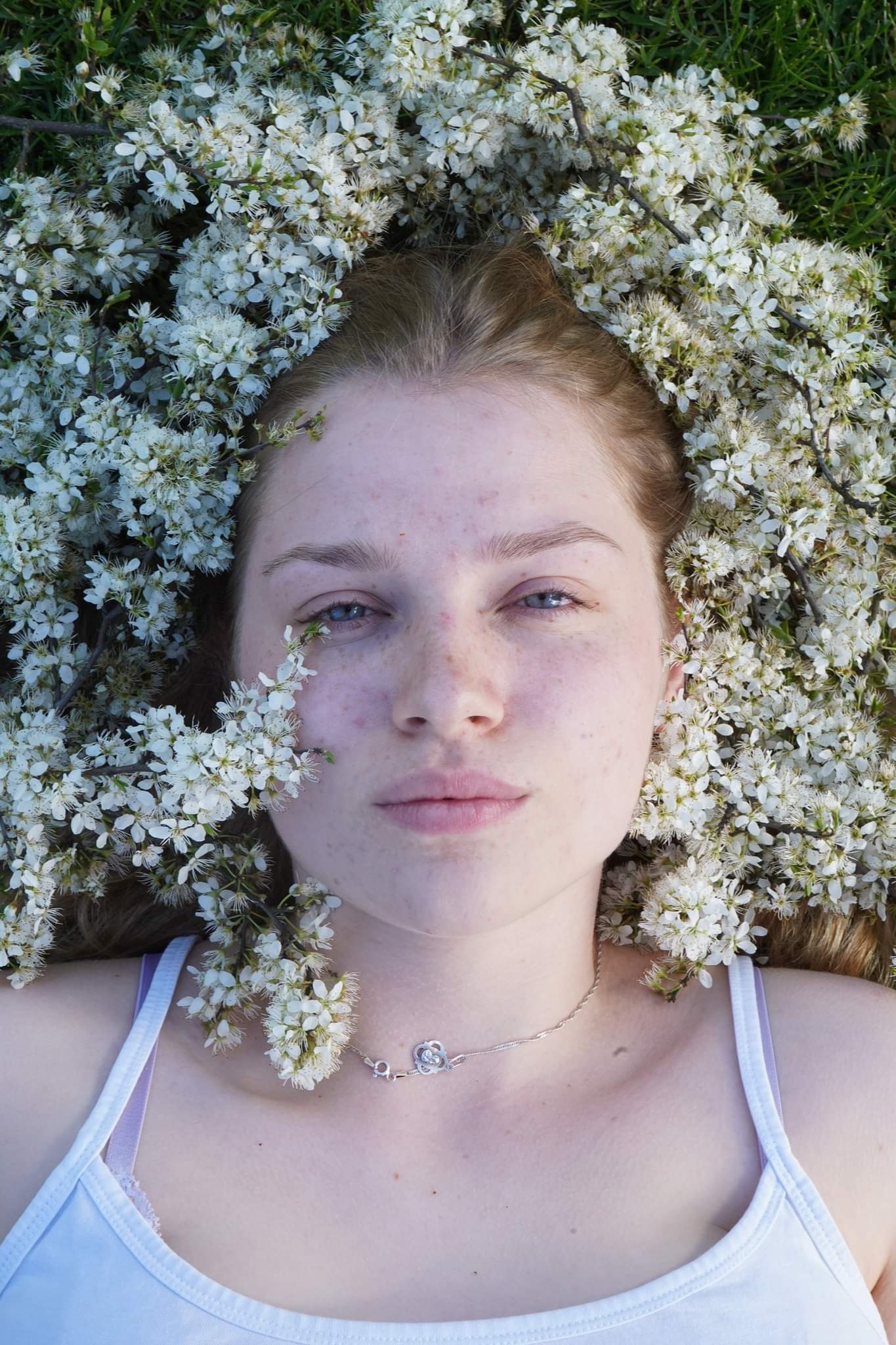 Portret młodej kobiety z rudymi włosami, leżącej na trawie, otoczonej białymi kwiatami, z delikatnym srebrnym naszyjnikiem.