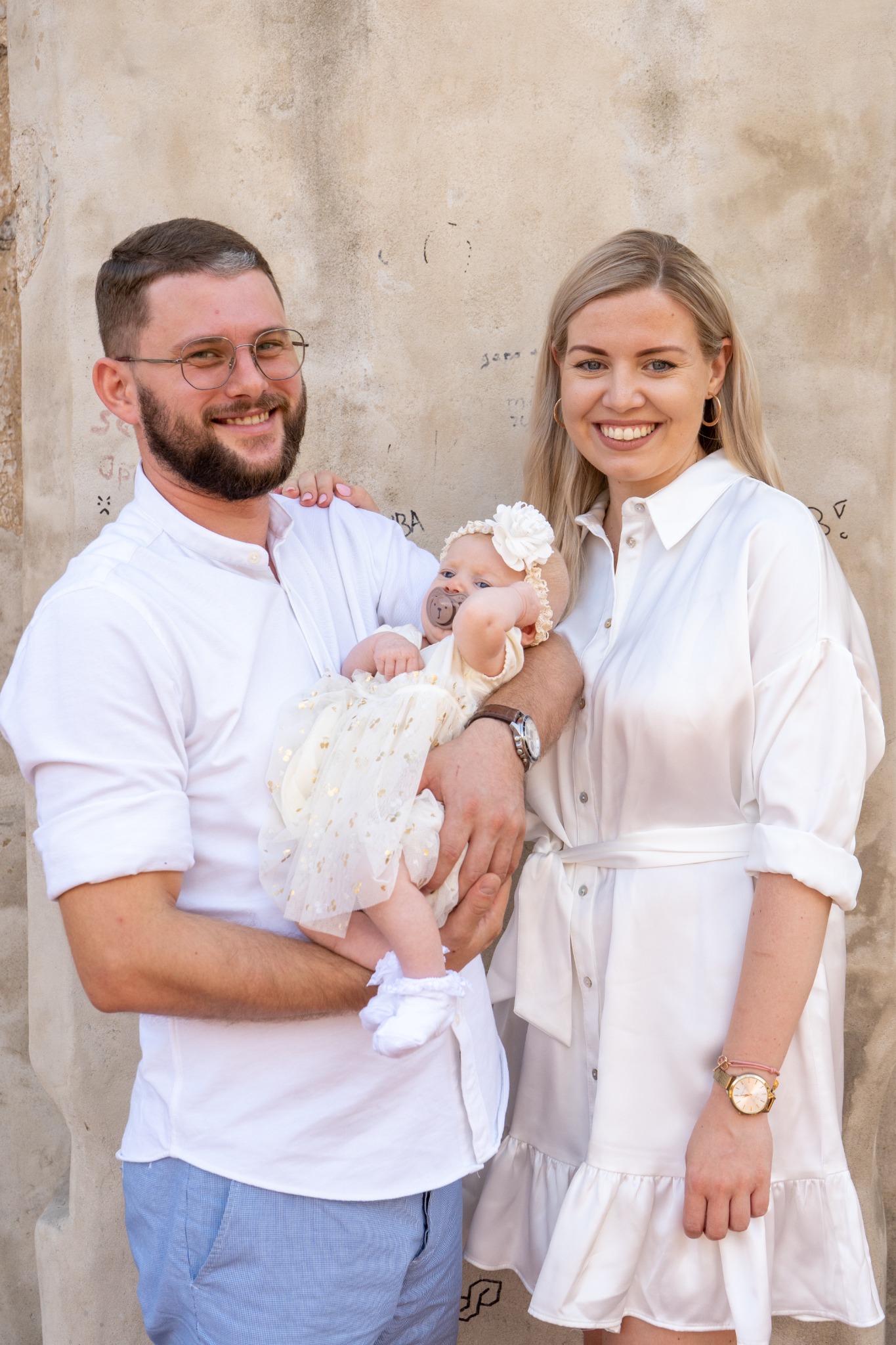 Rodzinne zdjęcie: Uśmiechnięci rodzice w białych strojach trzymają na rękach niemowlę w kremowej sukience z kokardą, na tle ściany z delikatnymi napisami.