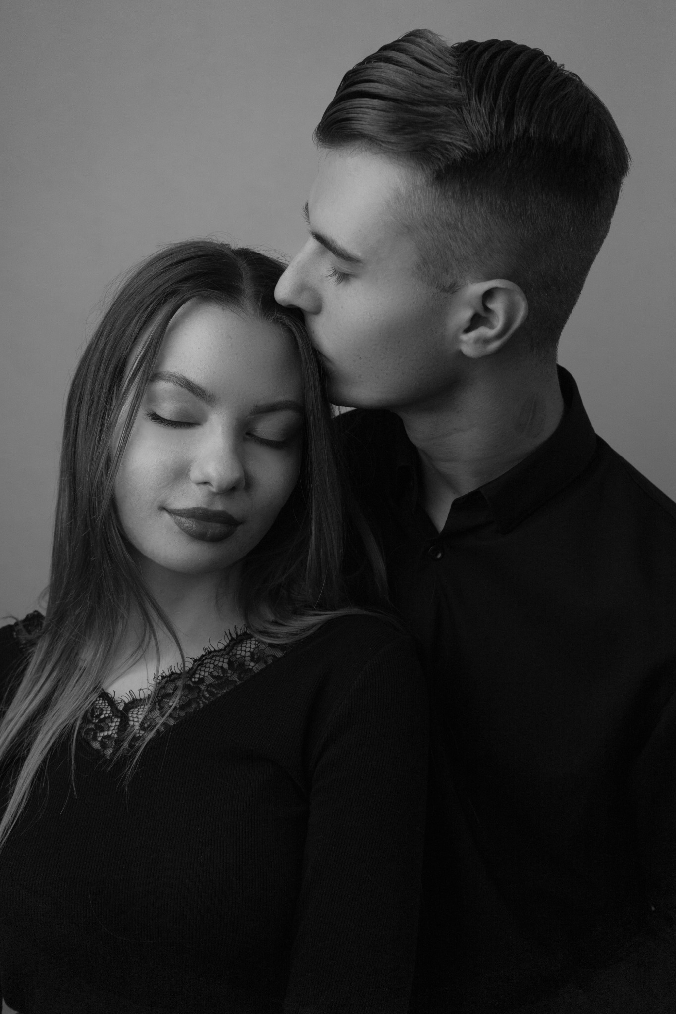 Czarno-białe zbliżenie: mężczyzna w czarnej koszuli całuje kobietę w czoło, oboje z zamkniętymi oczami na jednolitym tle.