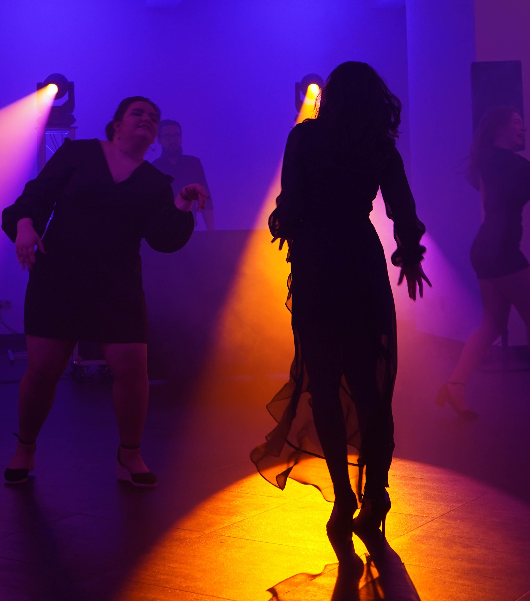 Sylwetki tańczących kobiet w sukienkach, oświetlone pomarańczowymi i fioletowymi światłami na parkiecie, z DJ-em w tle.