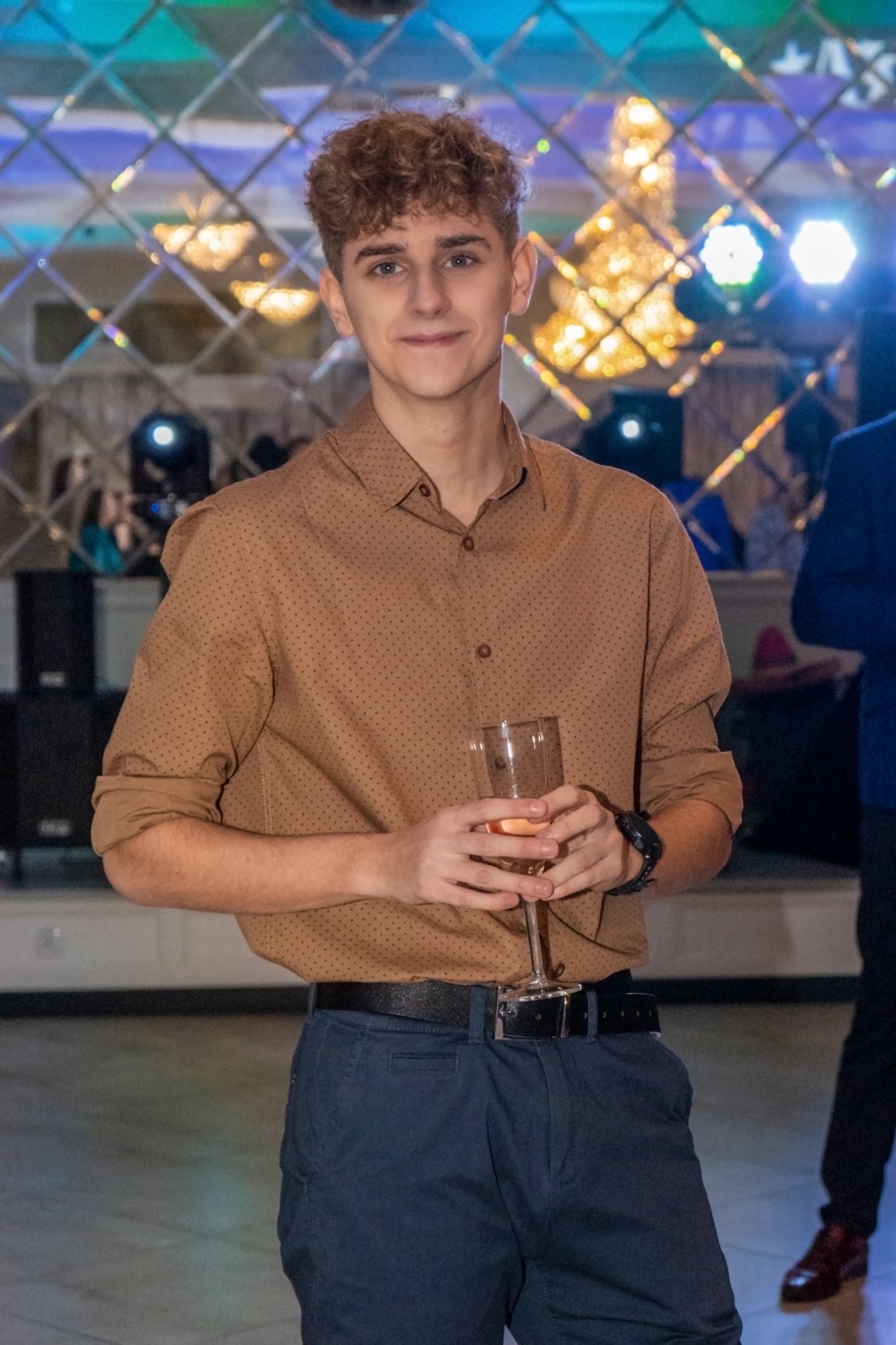 Młody mężczyzna w brązowej koszuli i granatowych spodniach, trzymający kieliszek z napojem na tle dekoracji lustrzanej i oświetlenia eventowego.