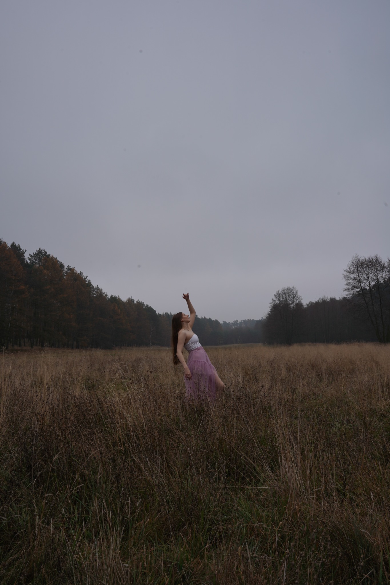 Kobieta w białym topie i tiulowej spódnicy pozuje na łące, wyciągając rękę ku pochmurnemu niebu, w tle las.